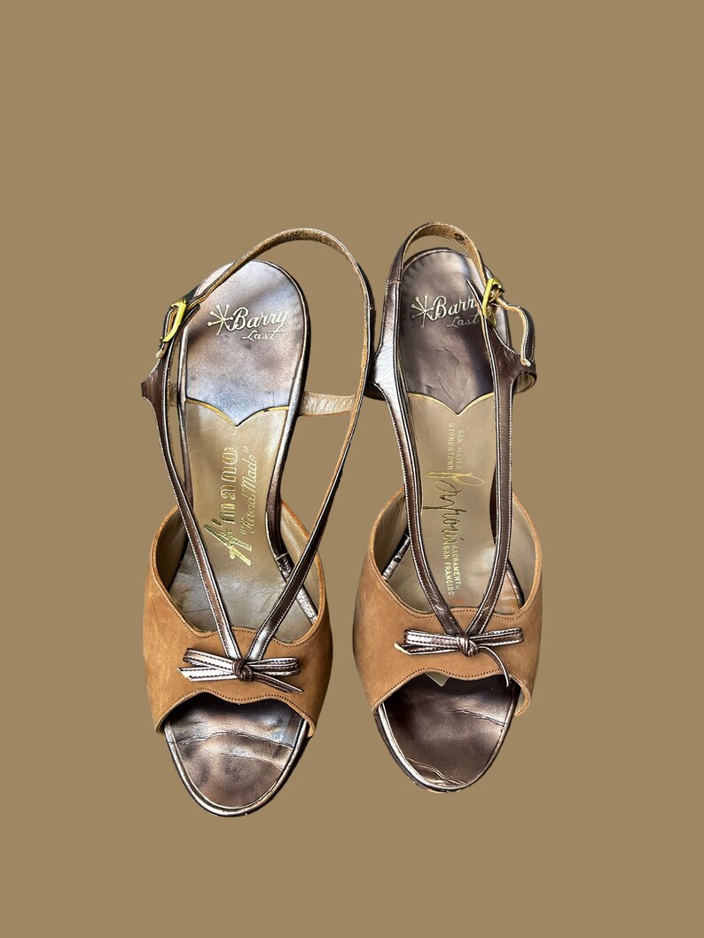 K&ouml;nnte beinhalten: Ein Paar braune Wildledersandalen mit Absatz, goldener Schnalle und Schleifendetail. Die Sandalen sind mit "Barry Last" und "Jeffrey California" gekennzeichnet.