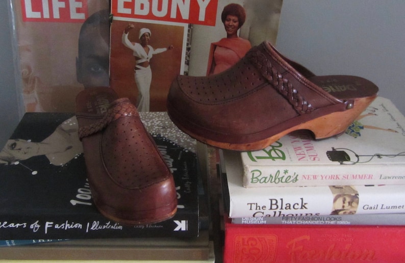 May include: A pair of brown leather clogs with a wooden sole. The clogs have a braided strap across the top and are sitting on top of a stack of books. The books have titles like "Barbie's New York Summer" and "The Black Californians".