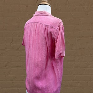 May include: A pink short-sleeved button-down shirt with a collar. The shirt is on a white mannequin against a brown brick wall.
