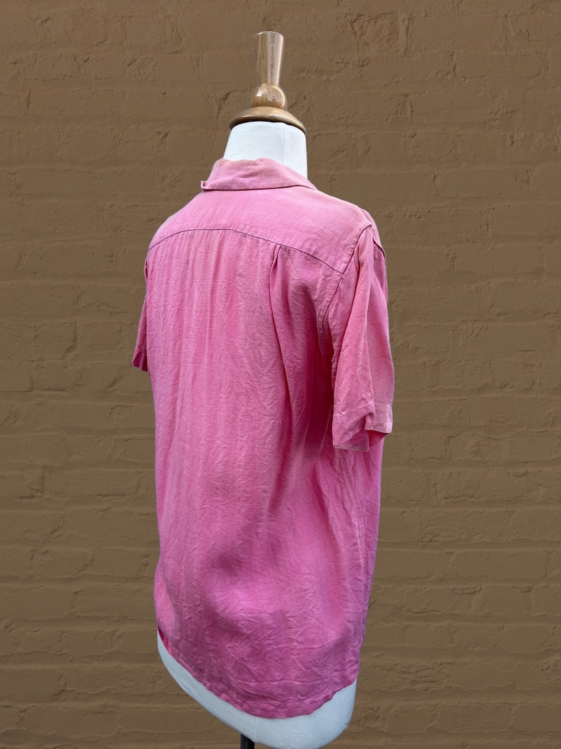 May include: A pink short-sleeved button-down shirt with a collar. The shirt is on a white mannequin against a brown brick wall.