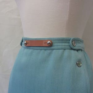 May include: A light blue skirt with a brown leather belt and silver buttons. The skirt has a wrap-around style and a single button closure.