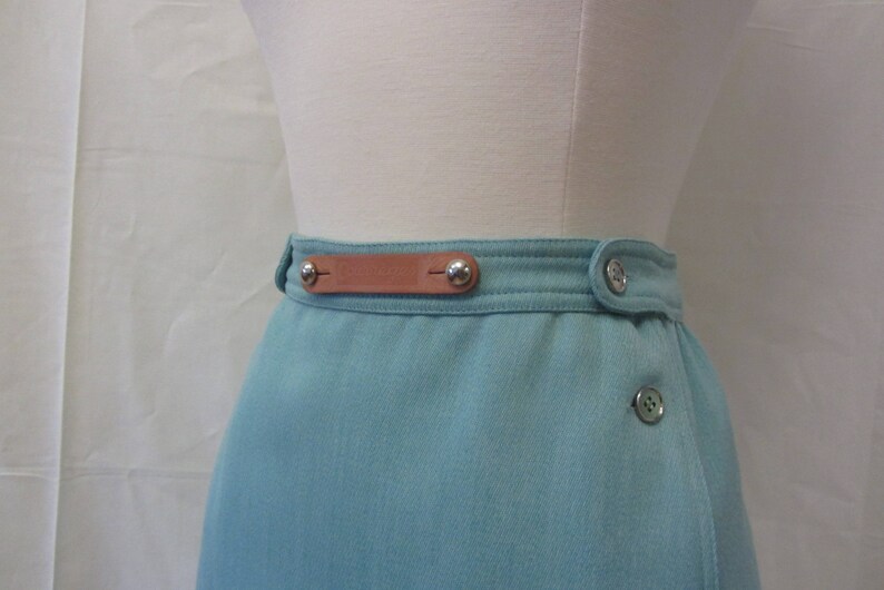 May include: A light blue skirt with a brown leather belt and silver buttons. The skirt has a wrap-around style and a single button closure.