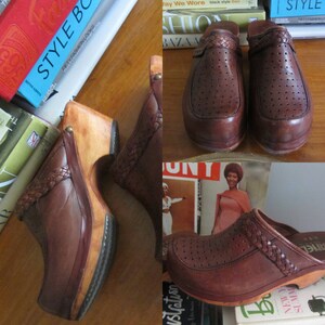 May include: A pair of brown leather clogs with a woven strap detail and a wooden platform sole. The clogs have a perforated design and are worn on a wooden floor.