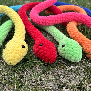 May include: A set of seven crocheted plush snakes in a rainbow of colors, including blue, green, yellow, orange, red, pink, and purple. The snakes are arranged in a semi-circle on a bed of green grass.