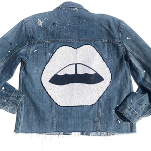 May include: A denim jacket with a large, sequined white lips design on the back. The jacket is a medium wash blue with paint splatters and a raw hem. The lips have a black outline and a black inner mouth.