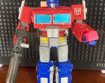 May include: A red and blue robot toy with a gun in one hand and a small red block in the other. The robot has a blue head with a silver face and a silver chest. The robot is standing on a wooden surface.