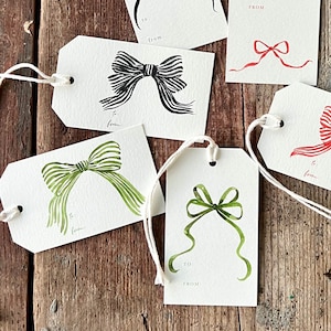 May include: White gift tags with watercolor bow illustrations in black, red, and green. Each tag has a string for attaching and space to write "To:" and "From:" on the front. The tags are arranged on a rustic wooden surface.
