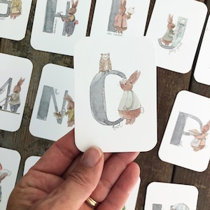 Może przedstawiać: Zestaw kart do nauki alfabetu z akwarelową ilustracją królika trzymającego inny przedmiot dla każdej litery. Litera C przedstawia królika trzymającego marchewkę.