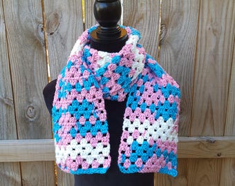 May include: A handmade crochet scarf in a granny square pattern. The scarf features a color block design with squares of light blue, pink, and white. The scarf is displayed on a black mannequin.