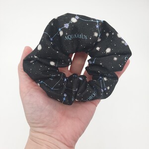 May include: Black scrunchie with a white background and a constellation print. The constellation is Aquarius and the text "Aquarius" is printed on the scrunchie.