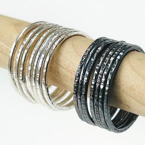 May include: A display of silver and black hammered metal bangles on a wooden dowel. The silver bangles are bright, while the black ones have a textured finish. The bangles are stacked, highlighting their design. The dowel is made of wood.