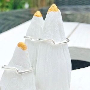 May include: Three silver rings with a wavy design are displayed on white cones with gold tips. The rings have a minimalist aesthetic. The background is a blurred outdoor scene. The rings are simple and elegant.