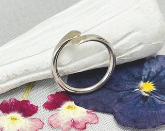 May include: A silver-colored ring with a unique open design, resting on a white stone. The ring is next to a deep purple flower with a yellow center and two smaller pink and white flowers. The background is a light gray fabric.