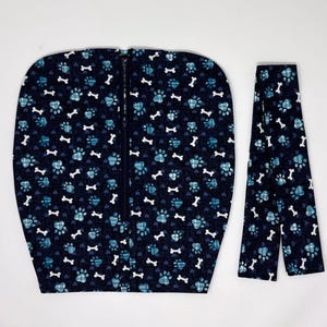 Puede incluir: Una funda de tela azul oscuro con un patrón repetitivo de huellas de patas a cuadros azul claro y huesos blancos, con una cremallera vertical central. Al lado hay una correa de tela larga y estrecha a juego con el mismo estampado.