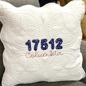 May include: White quilted pillow with a scalloped edge. The pillow has blue and red embroidered text that reads "17612 Columbia".