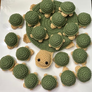 May include: A collection of crocheted green and beige turtle ornaments. The turtles are arranged in a circle around a larger, central turtle. The turtles are all different sizes and have a variety of crocheted details.