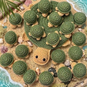 May include: A large crocheted turtle with a dark green shell and a light tan head, surrounded by many small crocheted turtles with dark green shells and light tan undersides, all arranged on a sandy surface with scattered seashells.