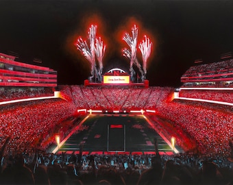 May include: A night view of a packed football stadium with a red sea of fans holding up red glow sticks. Fireworks explode in the night sky above the stadium. The scoreboard reads "Go Big Red!"