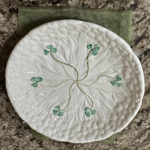 May include: A round white ceramic plate featuring a raised, textured floral pattern across the surface. The center of the plate is decorated with six hand-painted green shamrock motifs, each connected by thin, delicate stems radiating from the middle.