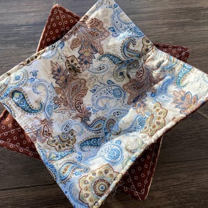 Puede incluir: Dos salvamanteles acolchados para cuencos. El superior tiene un fondo blanco con un estampado de cachemir en tonos azules, marrones y beige. El inferior es marrón con lunares blancos.