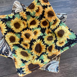 Puede incluir: Dos salvamanteles cuadrados de tela. Uno presenta un estampado de girasoles sobre un fondo negro con lunares blancos. El otro tiene un diseño de cachemira en tonos marrones, beige y azules. Los salvamanteles están apilados.