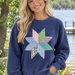 May include: A navy blue crewneck sweatshirt featuring a large, multi-patterned star appliqué on the chest. The star is constructed from various fabric patches, including blue gingham, floral, and solid green, with raw, frayed edges.