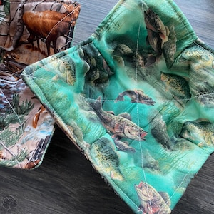 Puede incluir: Dos salvamanteles cuadrados acolchados. Uno presenta un patrón de camuflaje con ciervos y follaje. El otro tiene un fondo turquesa con un diseño de peces. Ambos están cosidos con hilo blanco.