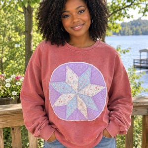 May include: A faded brick red crewneck sweatshirt featuring a large circular patch on the chest. The patch displays a quilted eight-pointed star design in shades of light blue, white, and lavender with a delicate floral pattern on the fabric.