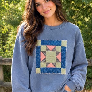 May include: A blue crewneck sweatshirt featuring a square quilt-style patch on the chest. The patch displays a central star pattern in shades of sage green, coral pink, and dark brown, framed by a dark blue border with a white circular print.