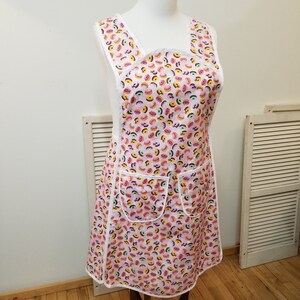 May include: A pink apron with a white trim and a pattern of colorful donuts. The apron has two pockets and a cross-back design. The apron is displayed on a mannequin.