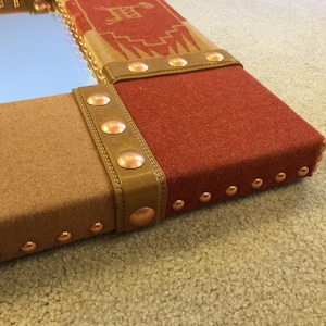 May include: A rectangular mirror with a brown and red fabric frame. The frame is decorated with copper-colored studs and leather straps.