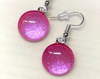 May include: A pair of pink glitter earrings with silver hooks. The earrings are round and have a gradient of pink glitter.
