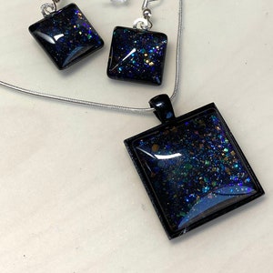 May include: A black necklace and earrings set with square pendants filled with iridescent glitter. The pendant and earrings are made of resin and have a black border.
