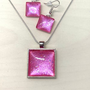 May include: A pink glitter necklace and earrings set. The necklace has a square pendant with a silver frame. The earrings are also square with silver frames.