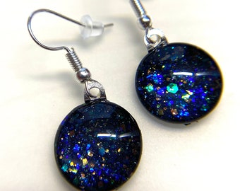 May include: A pair of silver dangle earrings with round black resin charms. The charms are filled with iridescent blue, green, and gold glitter.