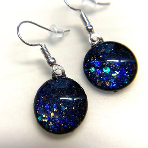May include: A pair of silver dangle earrings with round black resin charms. The charms are filled with iridescent blue, green, and gold glitter.