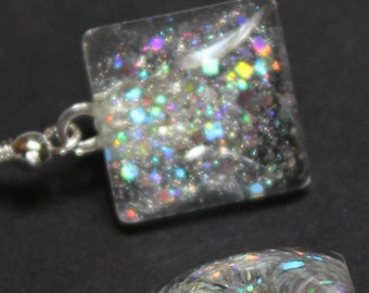 May include: A pair of square-shaped earrings with a clear, glitter-filled design. The glitter includes a mix of iridescent colors. The earrings have silver-colored fish hook ear wires.