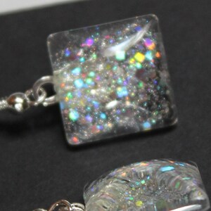 May include: A pair of square-shaped earrings with a clear, glitter-filled design. The glitter includes a mix of iridescent colors. The earrings have silver-colored fish hook ear wires.