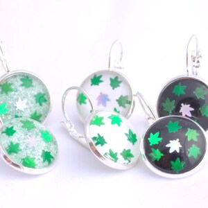 Puede incluir: Un conjunto de tres pares de pendientes colgantes de plata con diseños de hojas verdes. Los pendientes tienen cabujones redondos de vidrio con diferentes fondos: uno es blanco, otro es negro y otro es verde brillante.