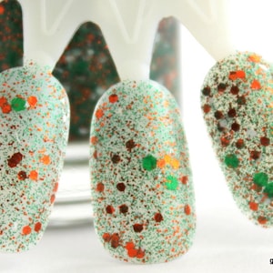May include: Close-up of three nail polish bottles with a white base and green and orange glitter. The bottles are on a white display stand.