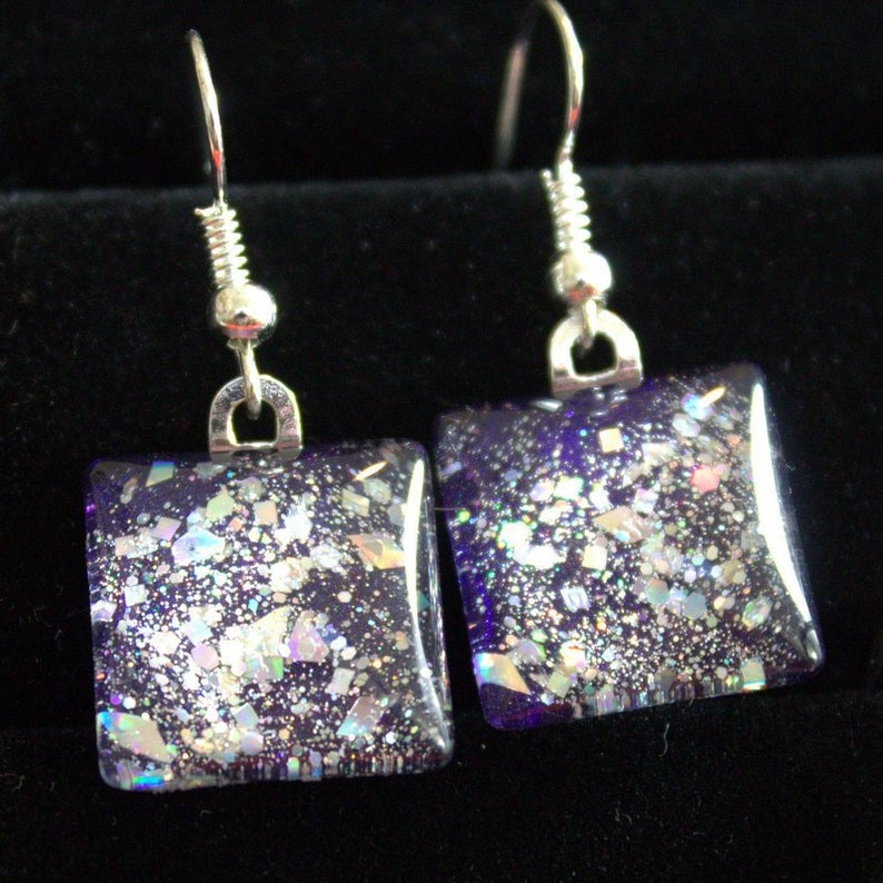 May include: A pair of square dangle earrings with silver hooks. The earrings are made of clear glass with iridescent glitter embedded in a purple base.