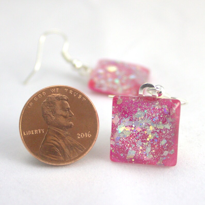 May include: Pink square earrings with iridescent glitter embedded in the resin. The earrings are shown next to a US penny for scale.