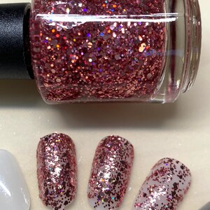 May include: A bottle of rose gold glitter nail polish with a clear glass bottle and a black cap. The polish is shown on four fingernails, each with a different amount of polish applied.