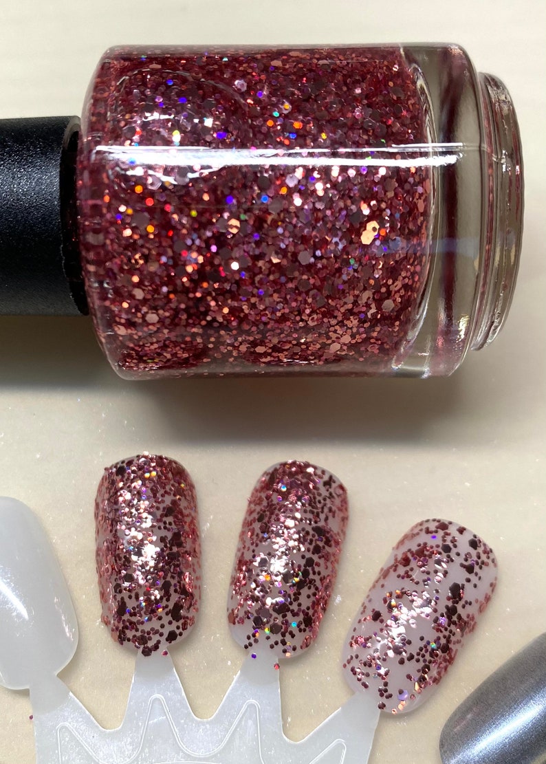 May include: A bottle of rose gold glitter nail polish with a clear glass bottle and a black cap. The polish is shown on four fingernails, each with a different amount of polish applied.