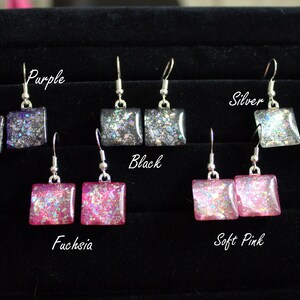 May include: A set of 6 pairs of dangle earrings with square-shaped glitter resin charms. The charms are in the colors purple, black, silver, fuchsia, and soft pink.