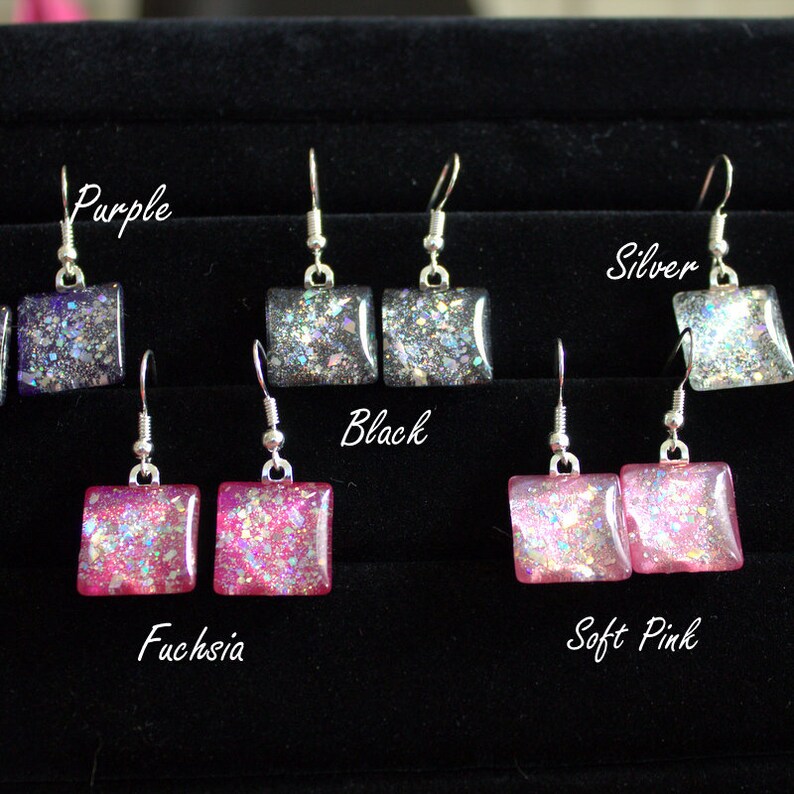May include: A set of 6 pairs of dangle earrings with square-shaped glitter resin charms. The charms are in the colors purple, black, silver, fuchsia, and soft pink.