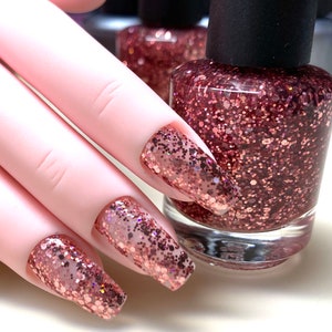 May include: Close-up of a hand with long, pink nails painted with a rose gold glitter nail polish. A bottle of the same rose gold glitter nail polish is in the background.
