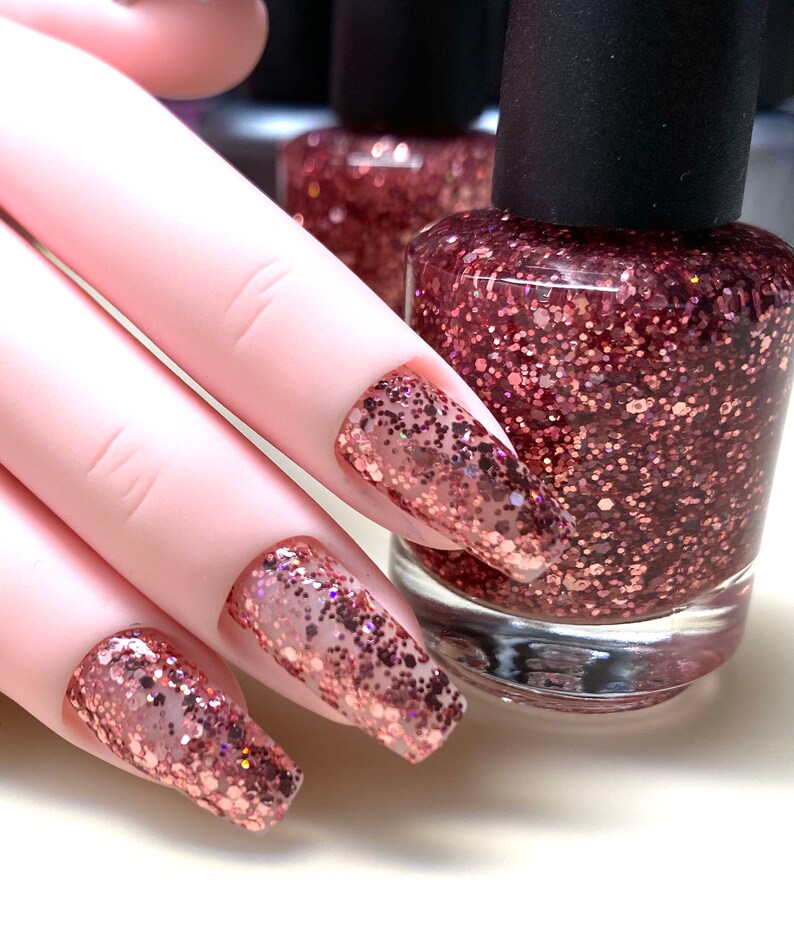 May include: Close-up of a hand with long, pink nails painted with a rose gold glitter nail polish. A bottle of the same rose gold glitter nail polish is in the background.