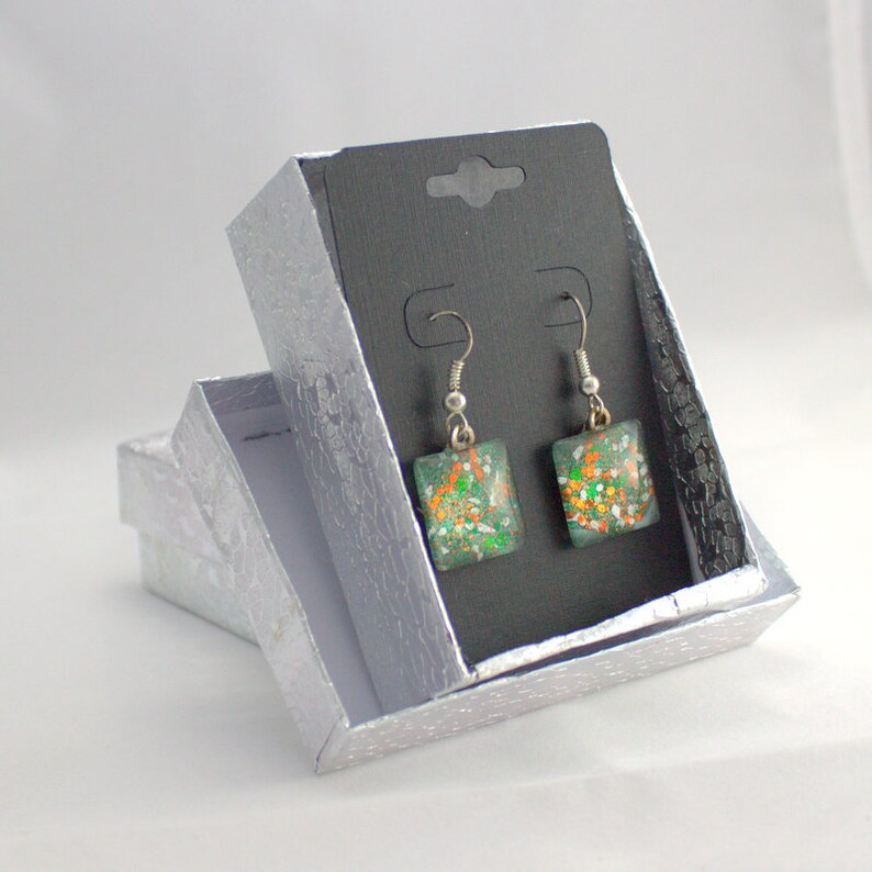 May include: A pair of square dangle earrings with green, orange, and white glitter embedded in clear resin. The earrings are silver and have a hook closure. The earrings are displayed in a silver gift box.