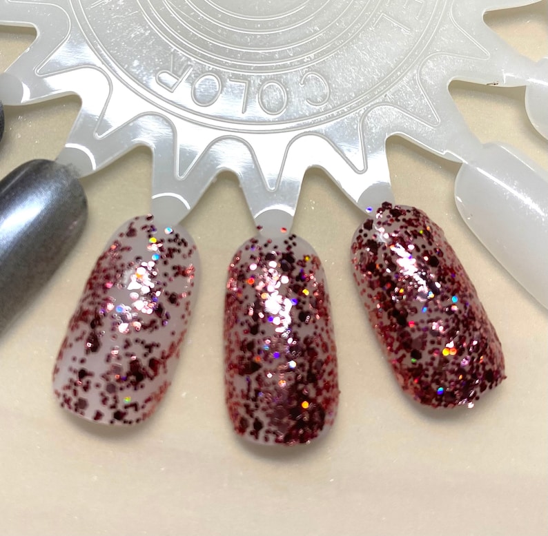 May include: Three nail polish swatches on a white color wheel. The swatches are a shimmery pink with red glitter.